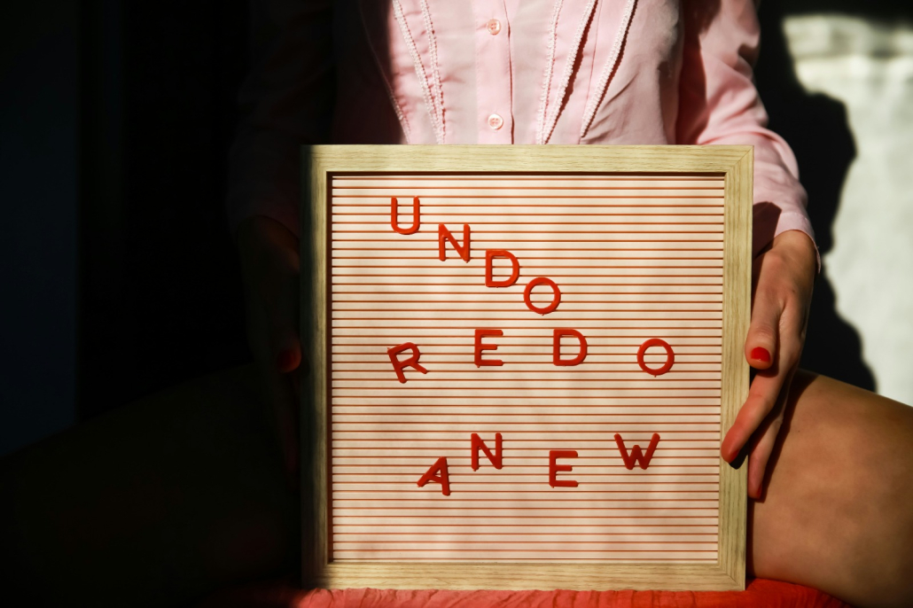 A person in a pale pink shirt holds a framed letterboard with red letters on a striped pink background. The words on the board, arranged diagonally and somewhat scattered, read: "UNDO," "REDO," and "ANEW." The person’s fingernails are painted red, and they are seated with the board resting on their lap, legs open, partially in shadow and light.